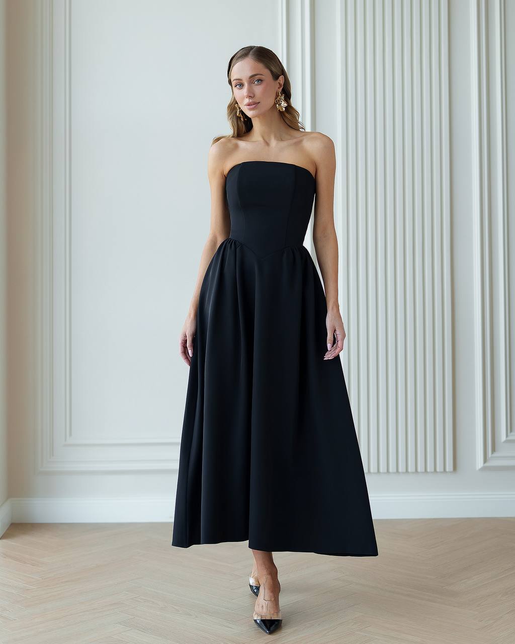Woman wearing a black strapless dress against a white paneled wall.