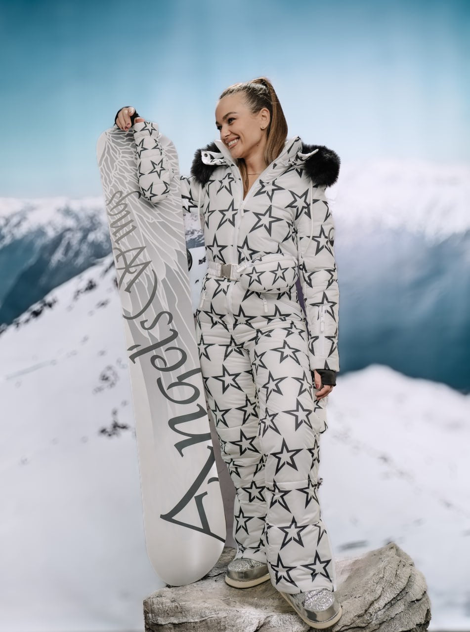 Person wearing a star-patterned snowsuit holding a snowboard with mountains in the background