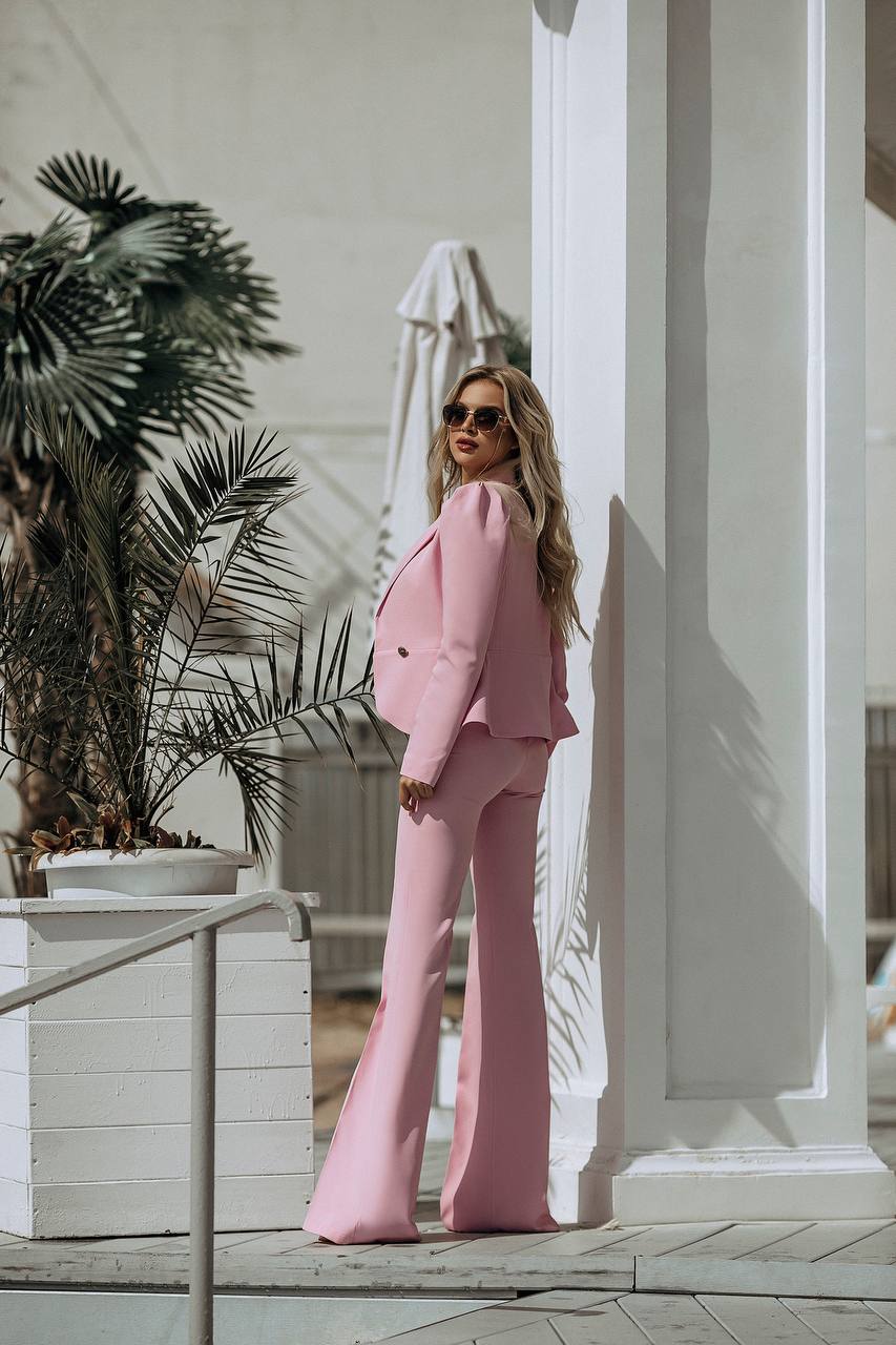 Woman in a pink suit standing outdoors with plants and a white building in the background