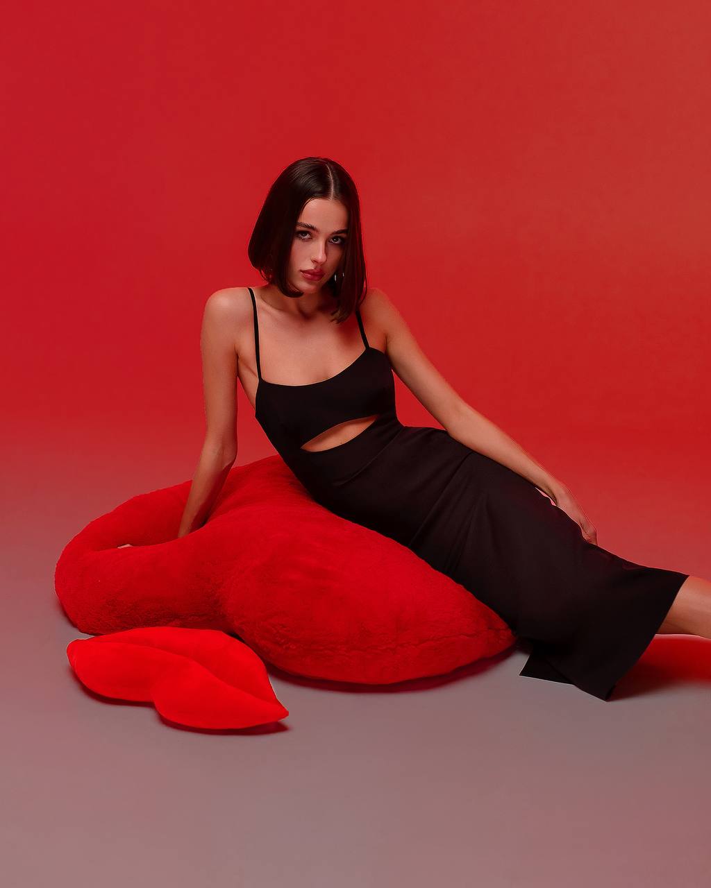 Woman in a black dress sitting on a red heart-shaped pillow against a red background