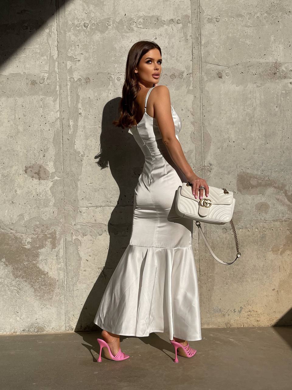 Woman in a white dress holding a white handbag against a concrete wall.