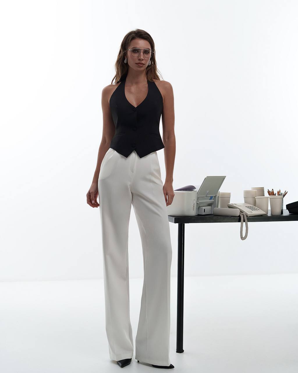 Woman in black top and white pants standing next to a desk with office supplies on a white background