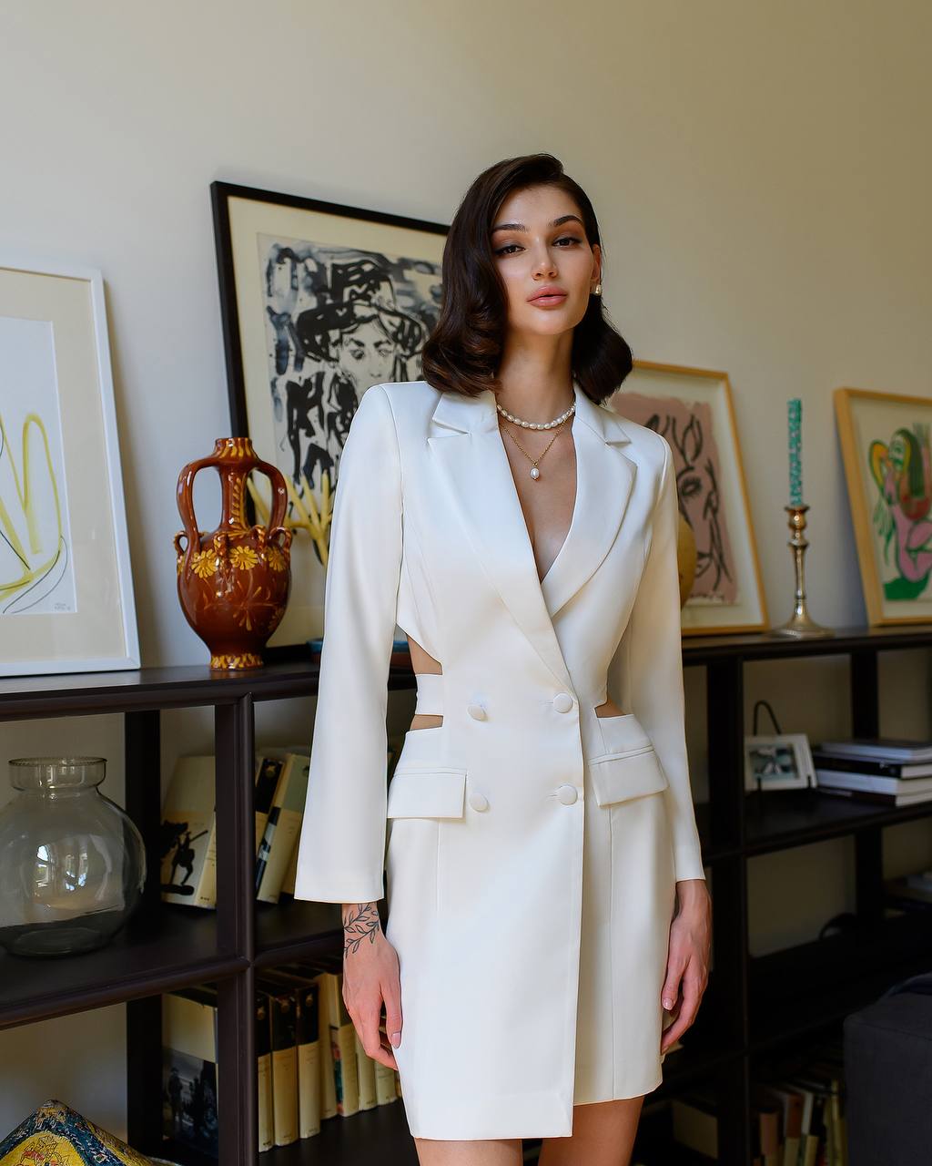 Woman in a white suit standing in a room with bookshelves and framed pictures.