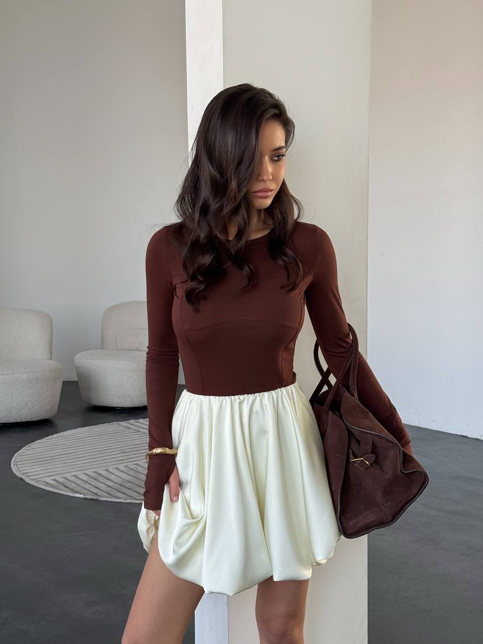 Woman in a brown top and white skirt holding a brown bag indoors.