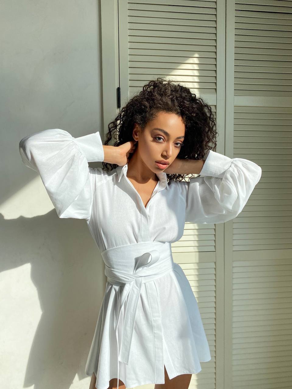 Woman wearing a white dress with wide sleeves in front of a window.