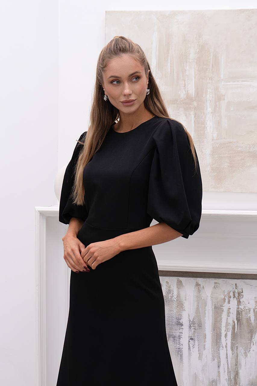 Woman wearing a black dress with puffed sleeves against a white wall.