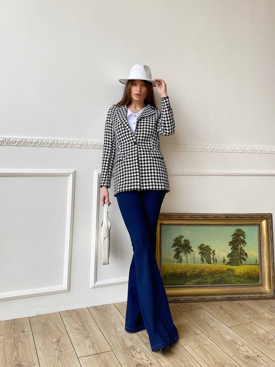 Woman in a checkered jacket and blue flared pants standing in a room with a painting on the wall.