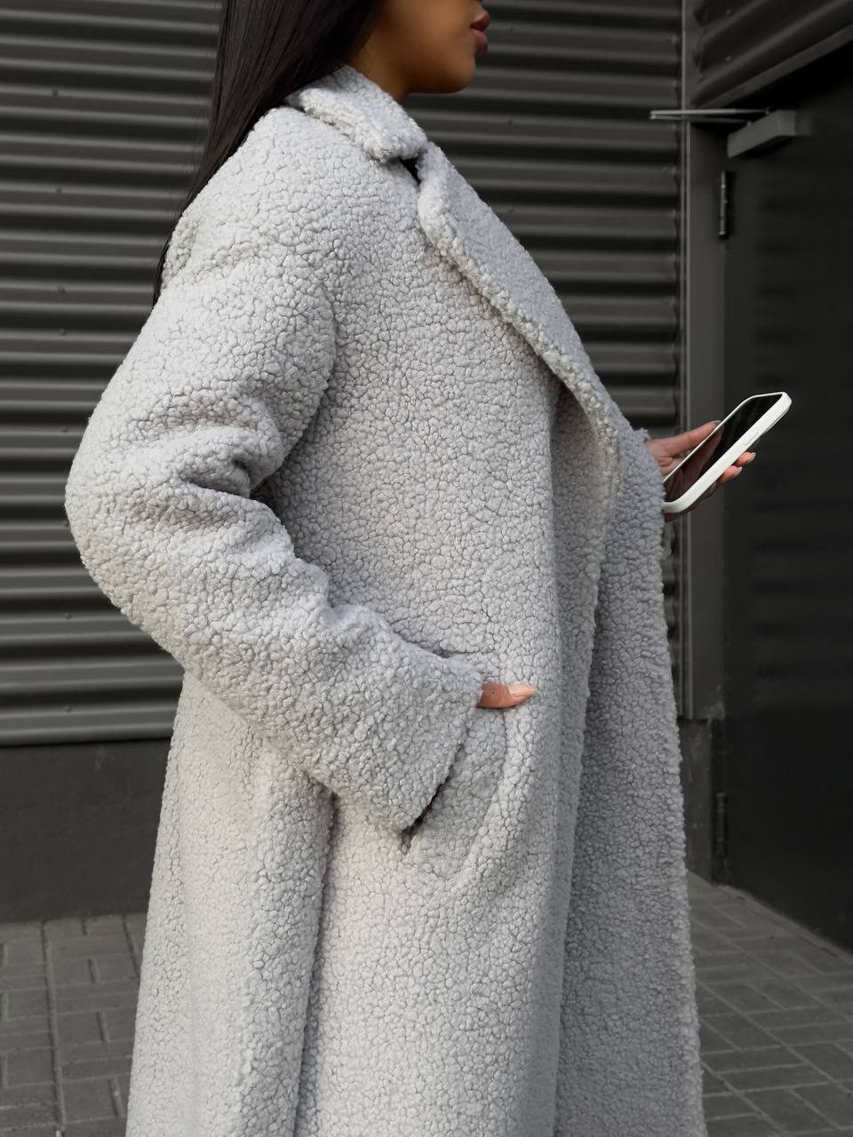 Person wearing a long light gray coat holding a phone against a dark background