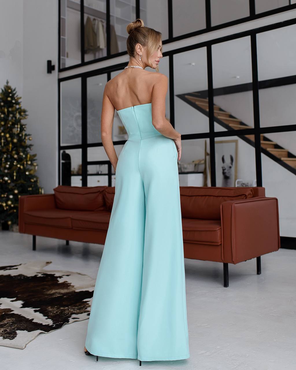 Woman in a light blue strapless jumpsuit standing in a modern living room.