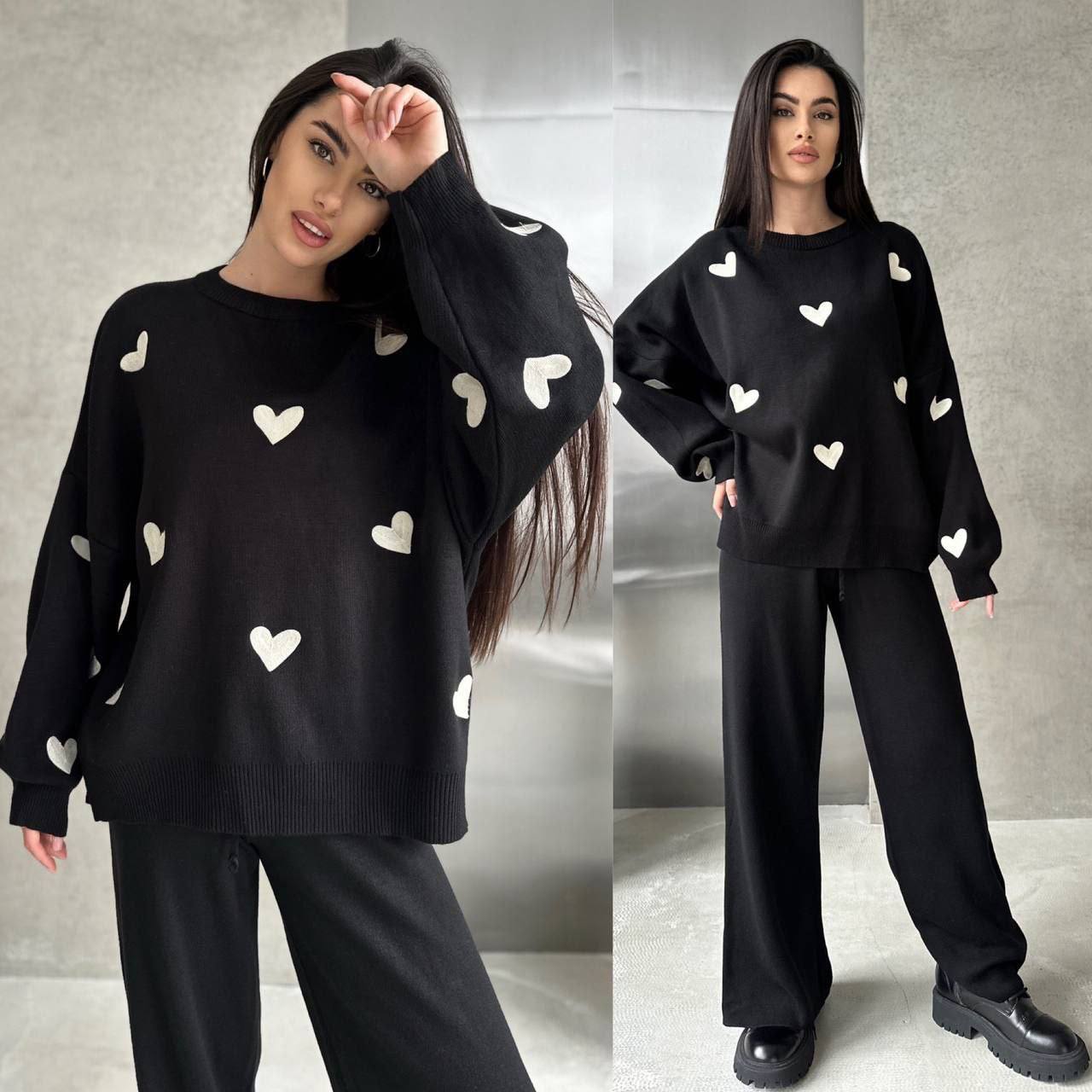Woman wearing a black sweater with white heart patterns and matching pants against a gray background.