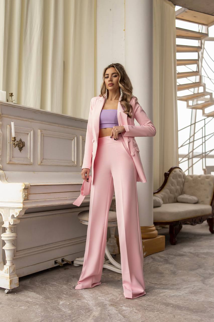 Woman in a pink suit standing in an elegant room with a piano and staircase.
