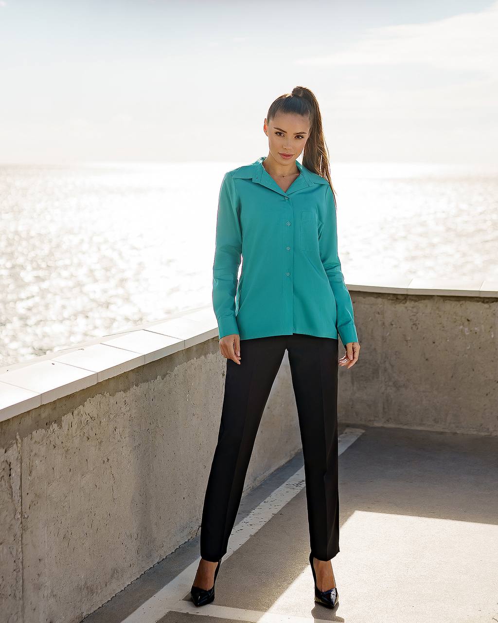 Woman wearing a teal jacket and black pants standing on a concrete ledge with a light background
