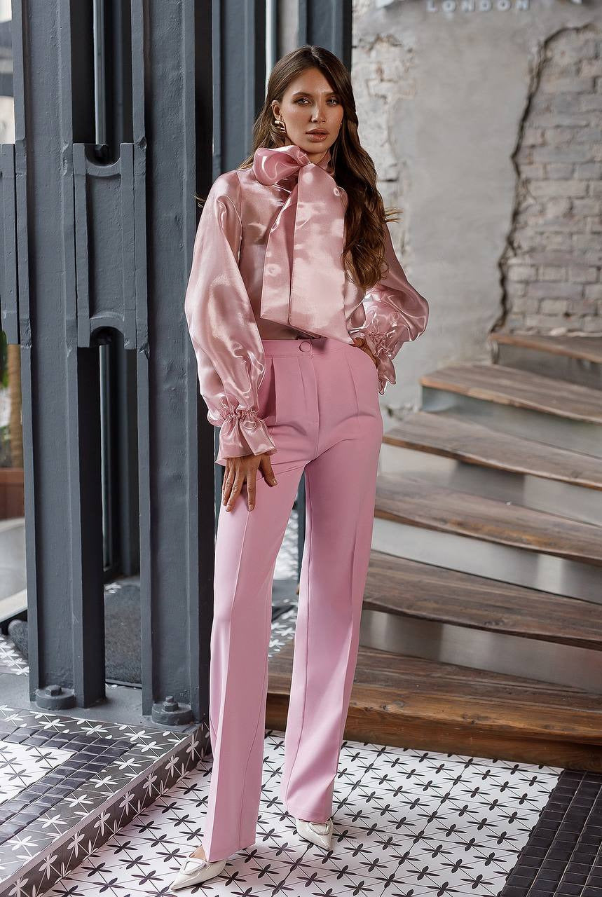 Woman in a pink outfit standing in an urban setting with a brick wall and metal structure.