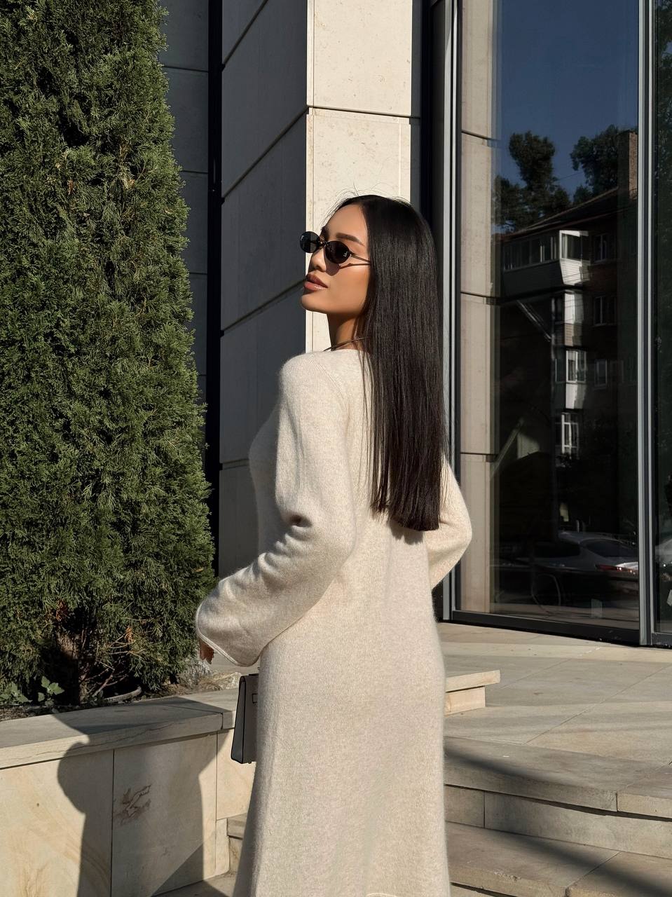 Woman in a long light-colored coat standing outdoors near a building with reflective windows.