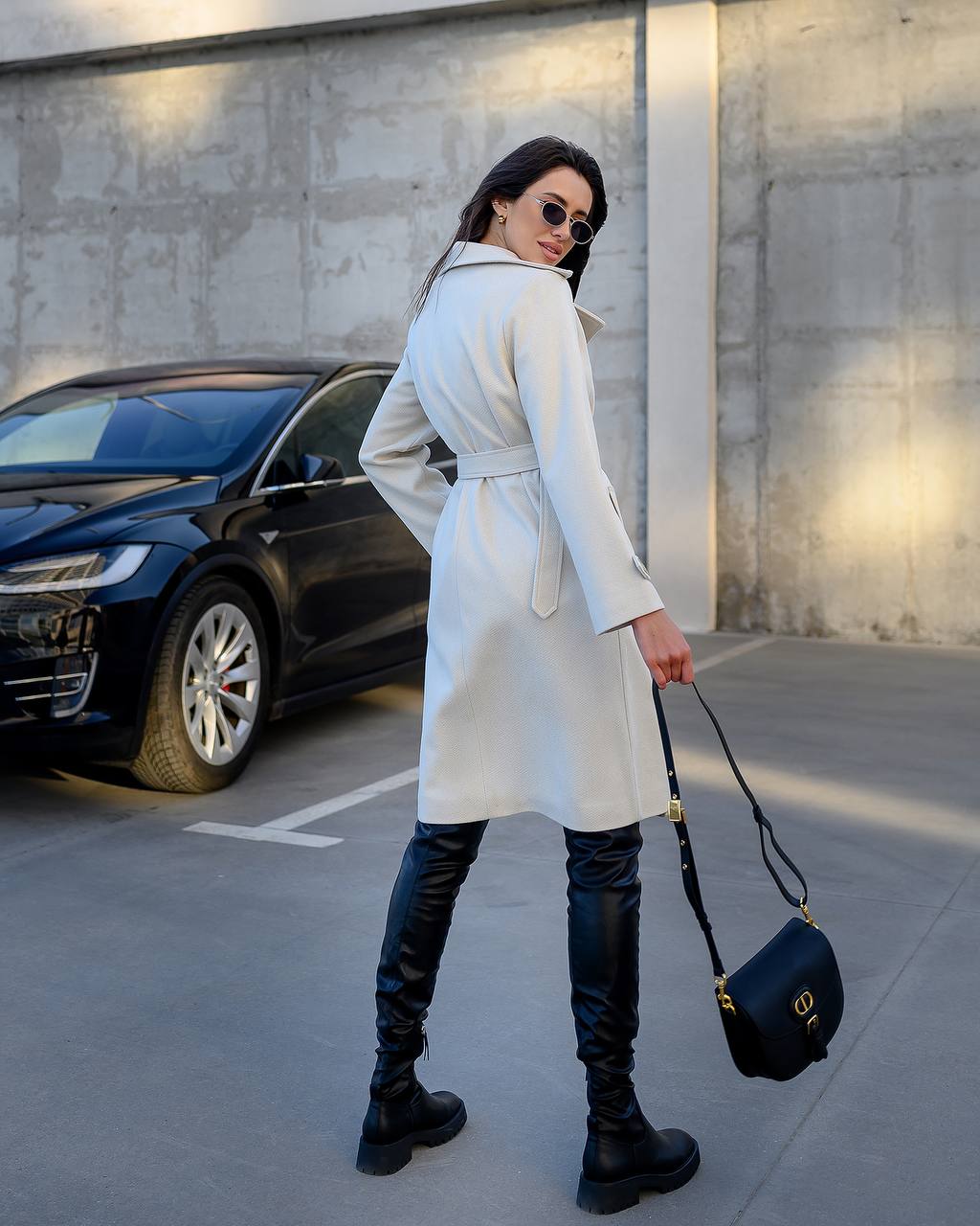 Woman in a white coat and black boots standing in a parking lot with a car in the background.