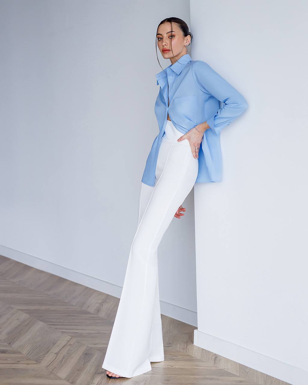 Person wearing a light blue shirt and white pants standing against a white wall.