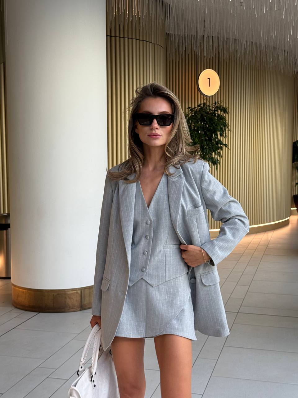 Woman in a light gray suit standing indoors with decorative elements in the background