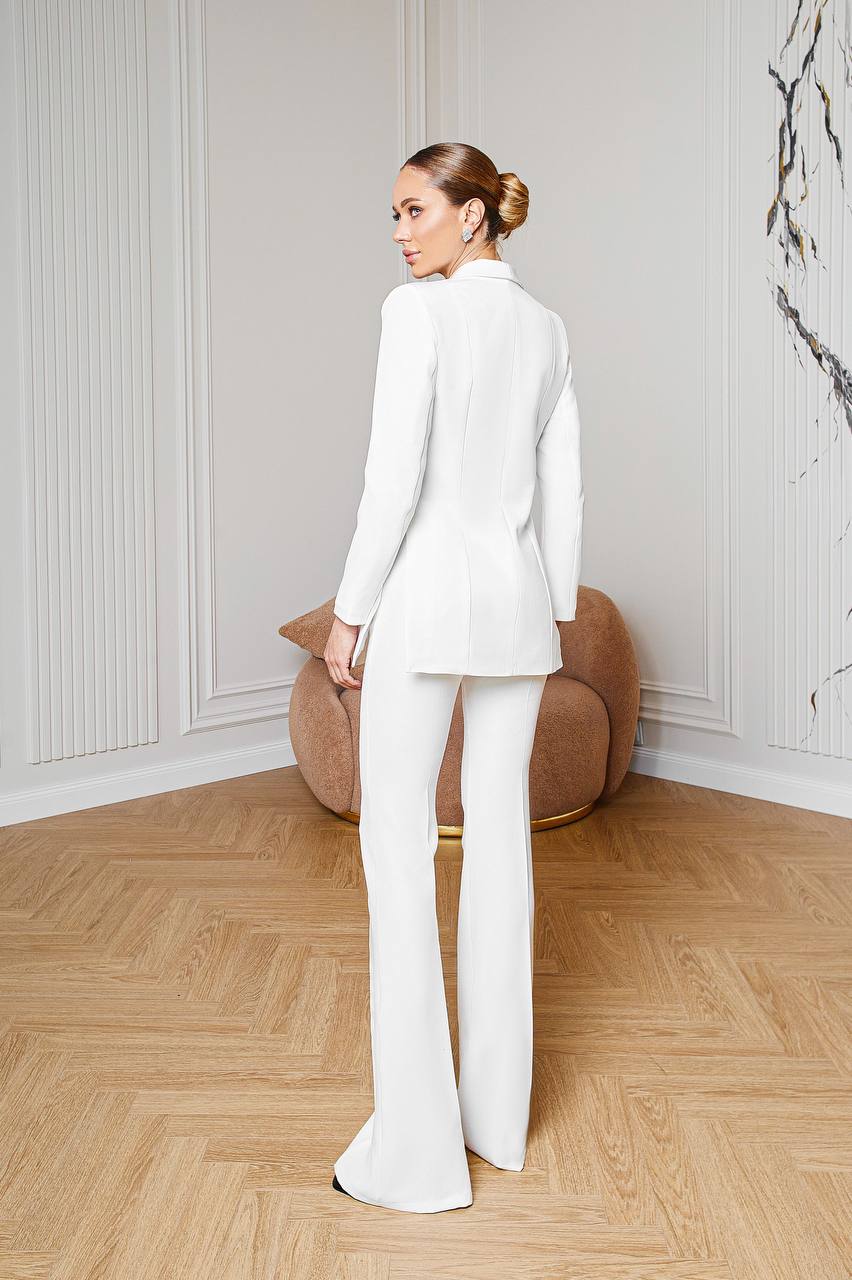 Woman in a white suit standing in a room with wooden flooring and a decorative wall.