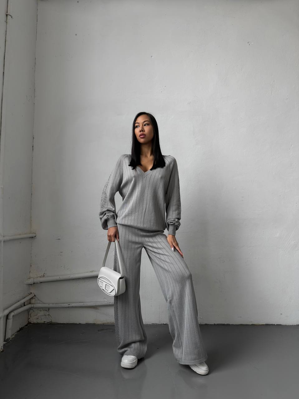 Woman in gray outfit holding a white bag against a plain white wall