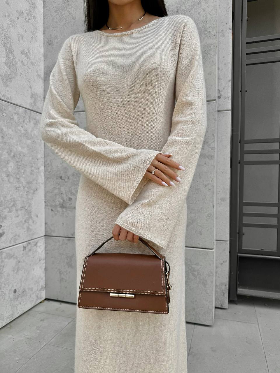 Person wearing a beige sweater dress holding a brown handbag against a neutral background