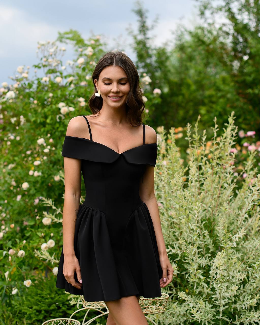 Woman wearing a black dress standing in a garden with greenery and flowers.