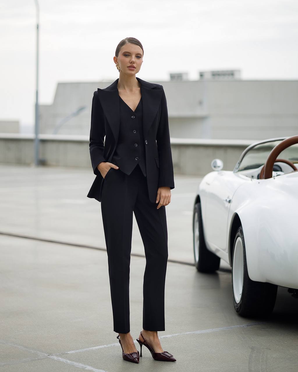 Woman in a black suit standing next to a white car on a rooftop.