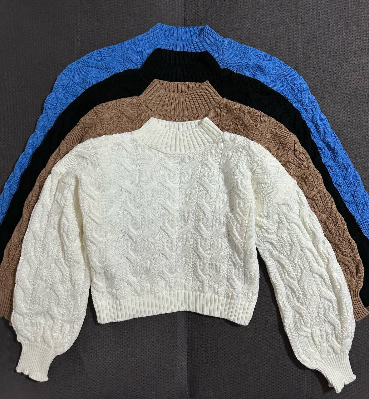 Stack of cable knit sweaters in white, brown, and blue on a dark background
