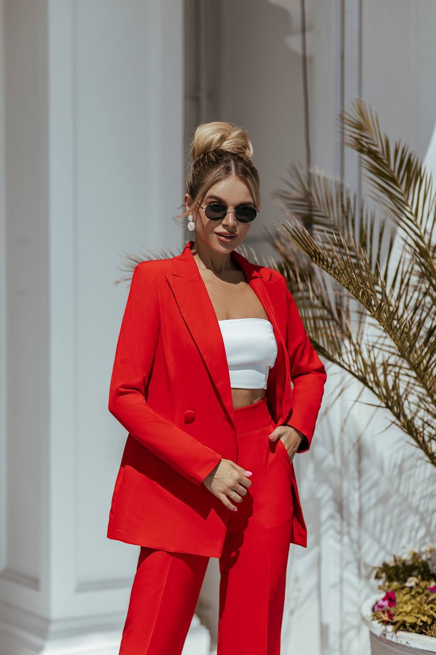Woman in a red suit standing against a white wall with plants.