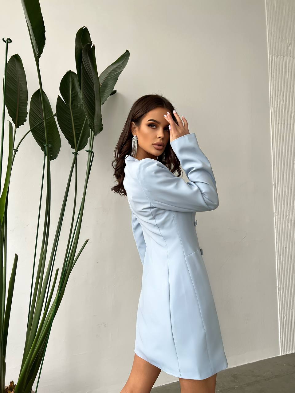 Woman in a light blue dress standing next to a plant against a plain wall