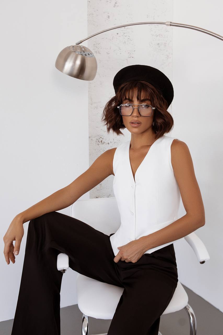 Woman in a white sleeveless top and black pants sitting on a white chair with a white wall and silver lamp in the background.