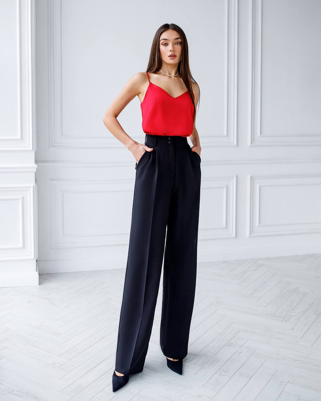 Woman wearing a red top and black wide-leg pants in a white room.