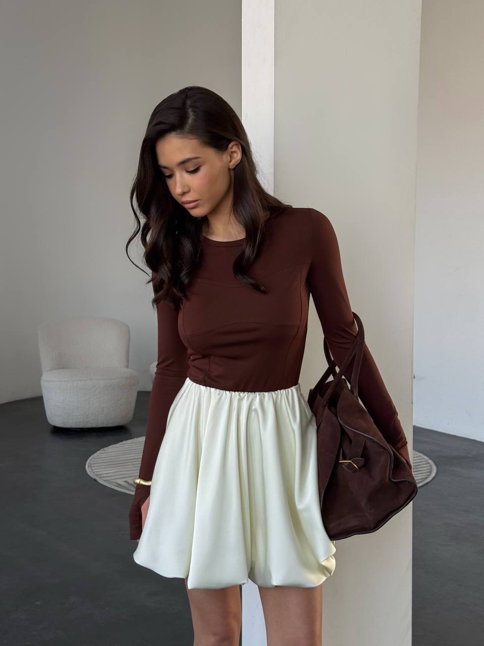 Woman wearing a brown long-sleeve top and white skirt, holding a brown bag indoors.