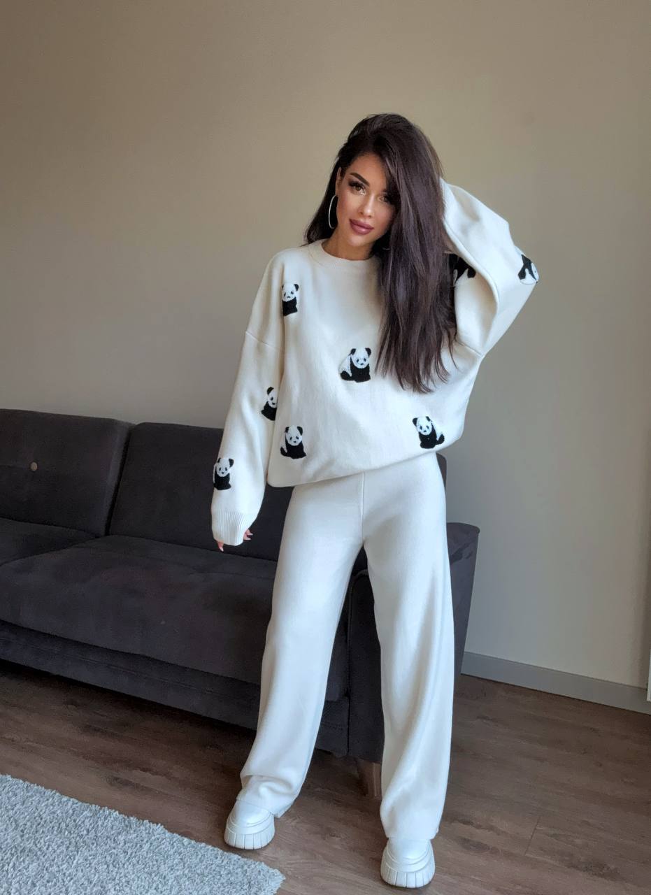 Woman wearing a white sweater with panda patterns and white pants in a room with a gray sofa.