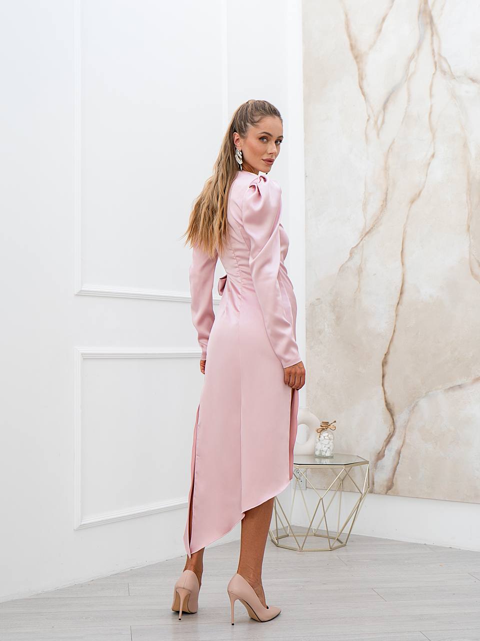 Woman in a pink dress standing in a room with marble wall and glass table.
