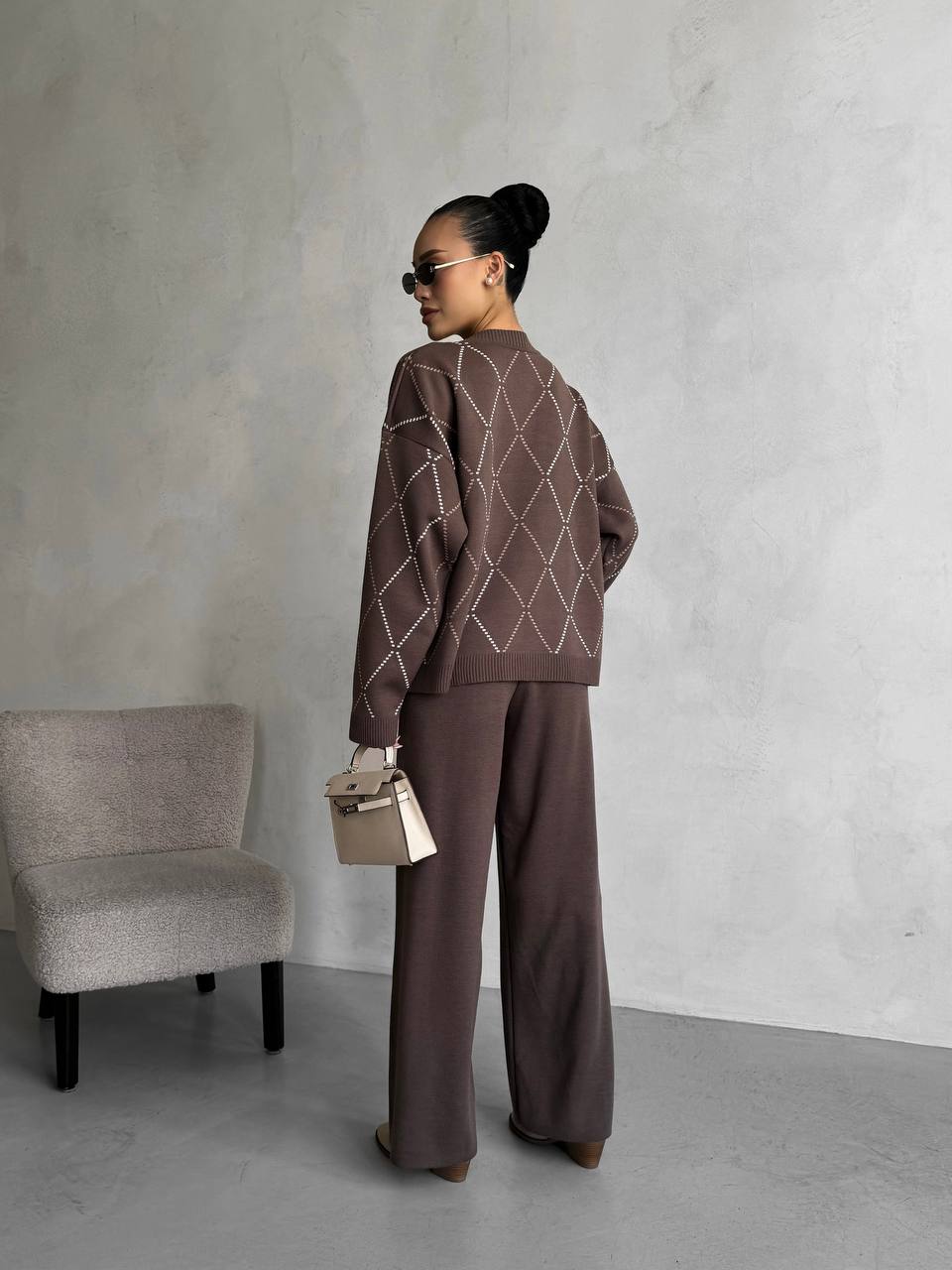 Person wearing a brown outfit with a geometric pattern, standing against a plain wall with a chair.