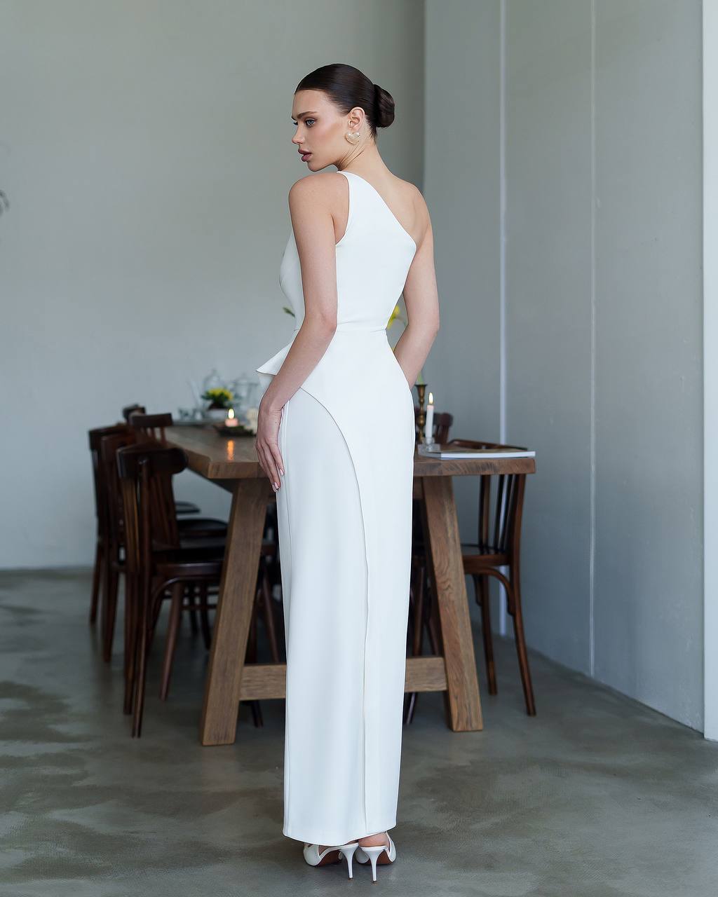 Woman in a white dress standing in a room with wooden furniture and a neutral color palette.