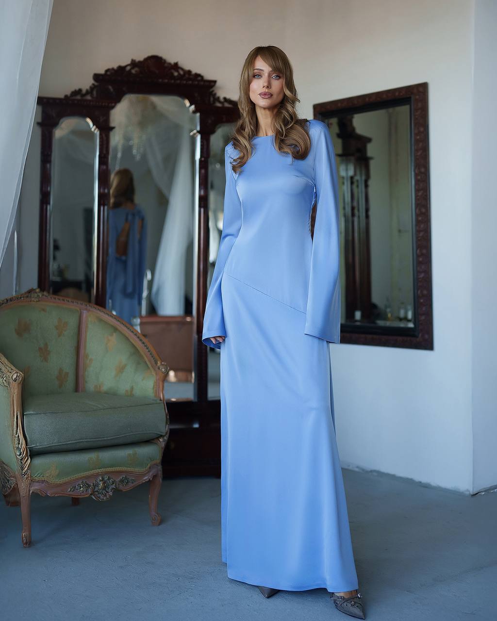 Woman in a long light blue dress standing in a room with mirrors and a chair.