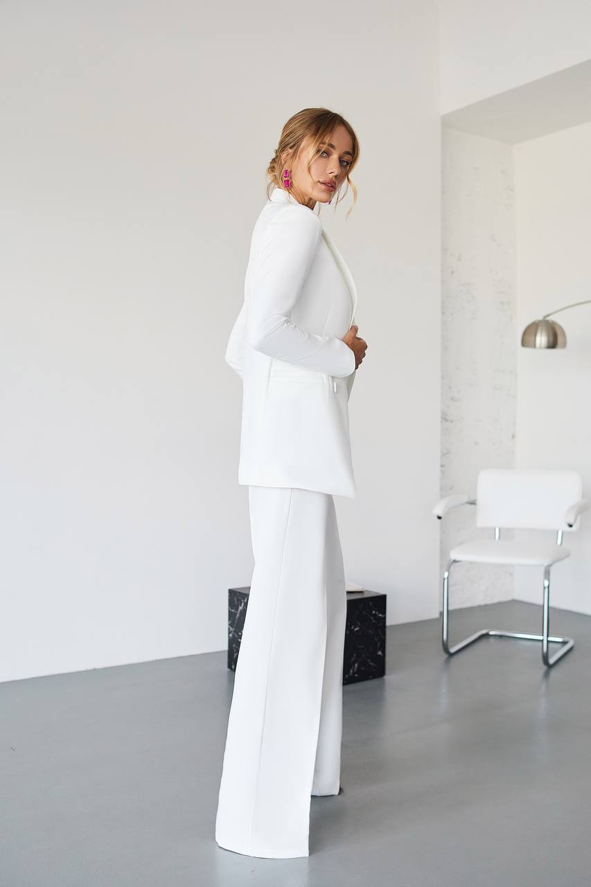Woman in a white suit standing in a minimalistic room with a white chair and lamp.