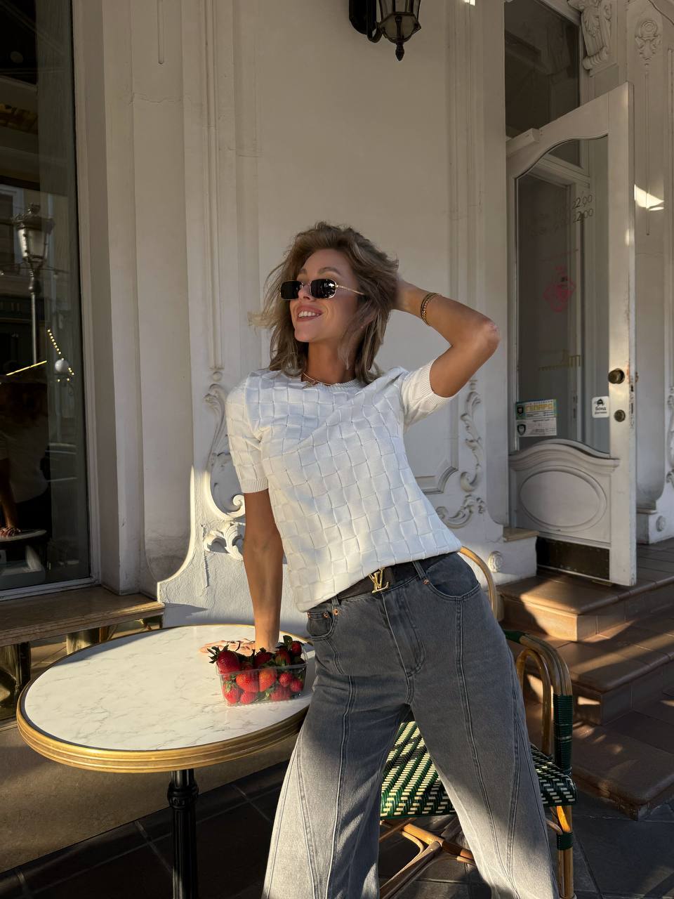 Woman in a white top and jeans standing outdoors near a table with a basket of flowers.