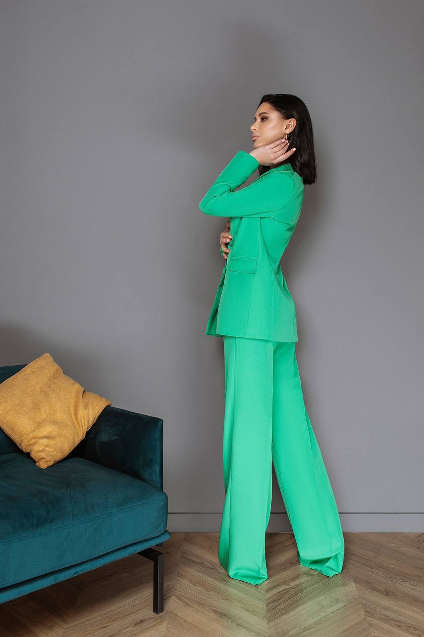Woman in a green suit standing next to a teal sofa with a yellow pillow against a gray wall.