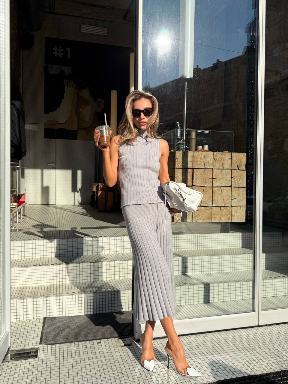 Woman in a gray dress standing outside a building holding a drink.