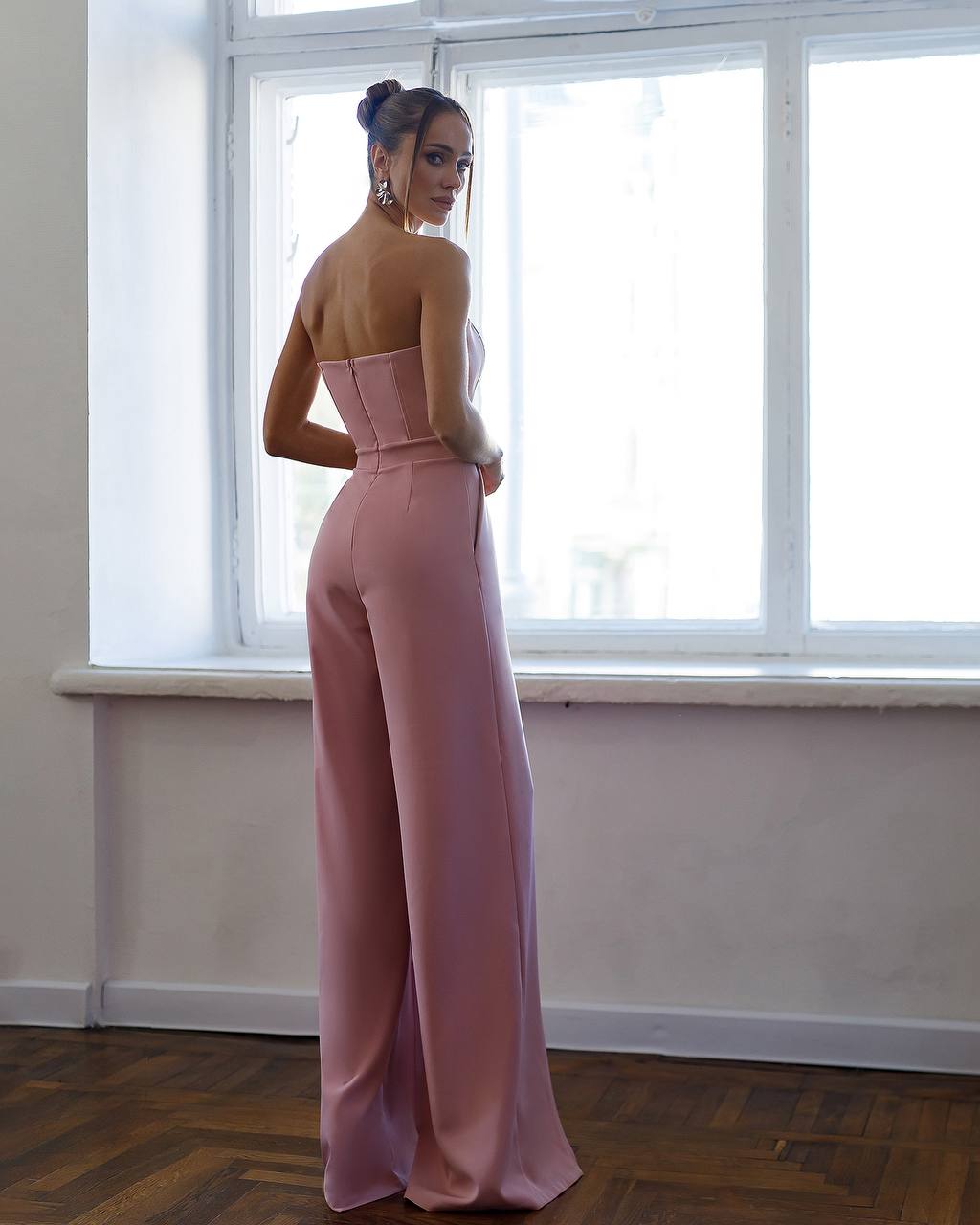 Woman wearing a pink strapless jumpsuit standing in front of a window.