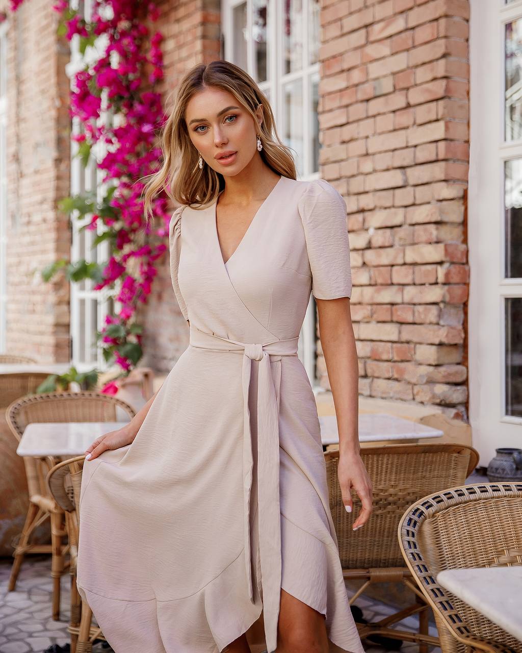 Woman in a light pink dress standing outdoors with floral decorations and a brick wall background.
