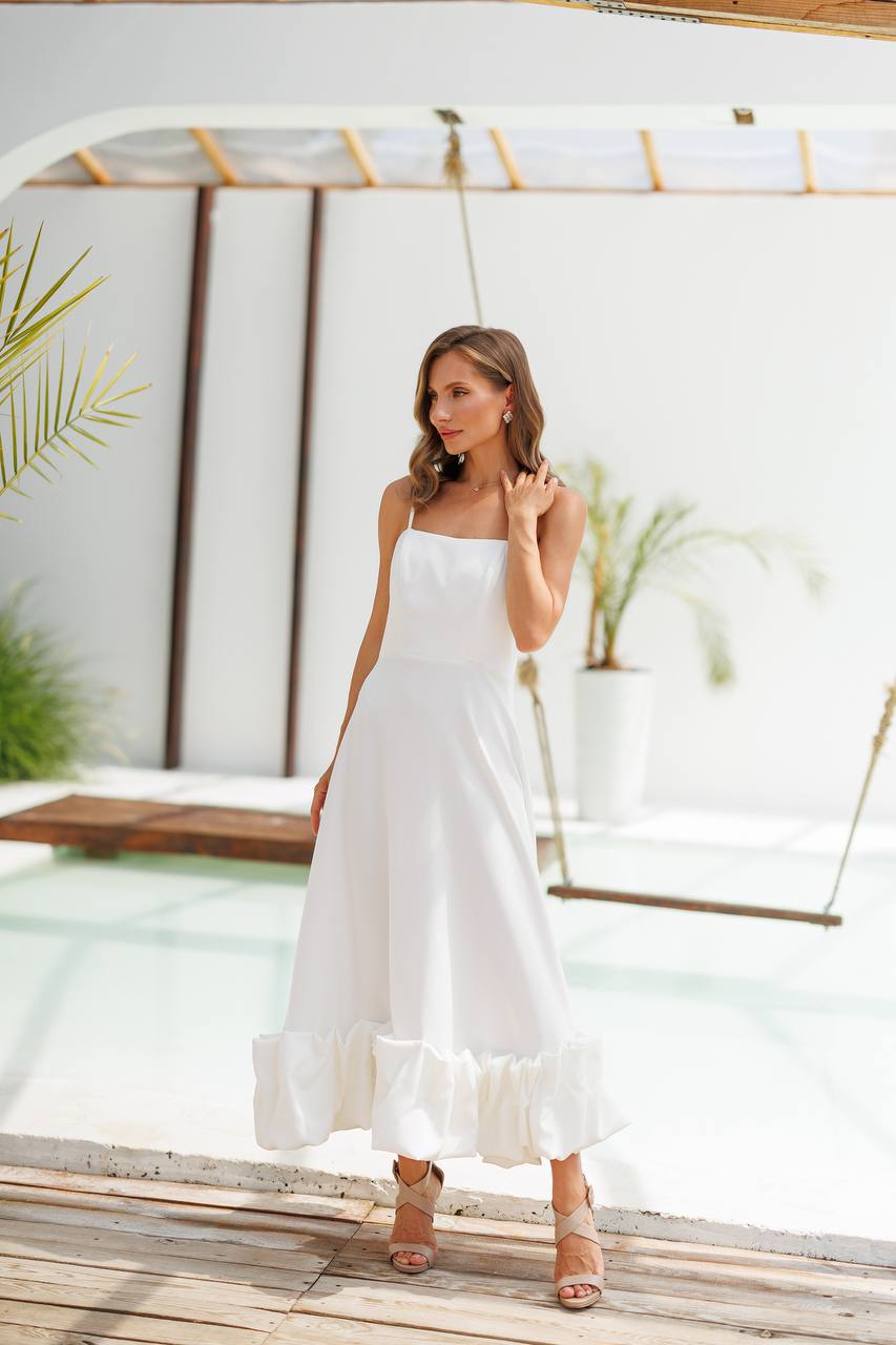 Woman in a white dress standing on a wooden deck with a white wall and plants in the background