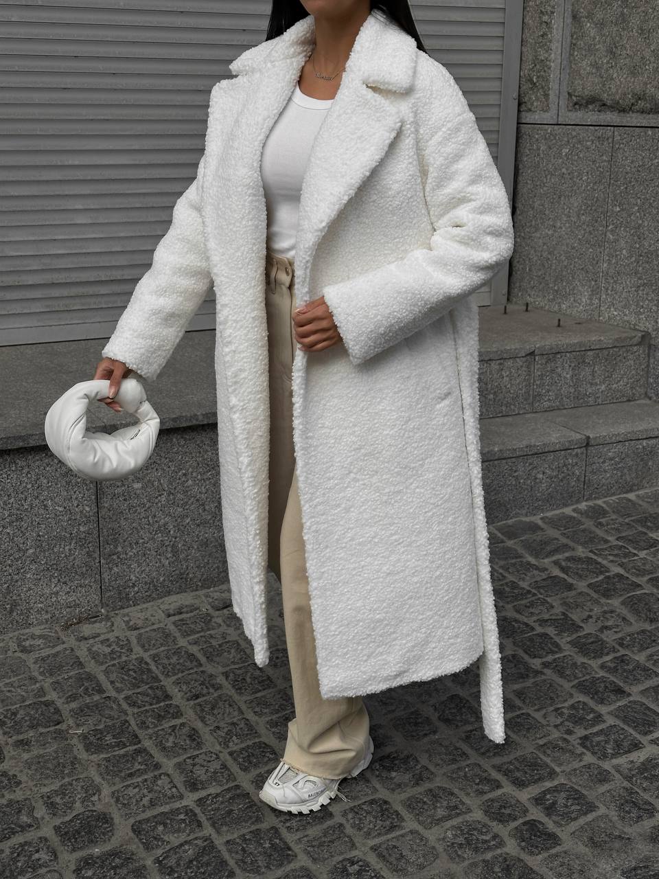 Person wearing a long white coat with a textured texture, standing on a sidewalk.