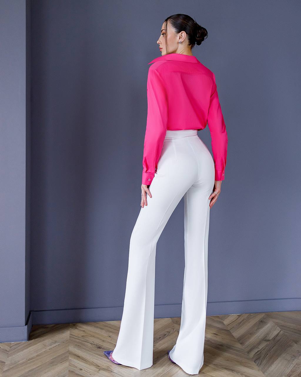 Woman wearing a bright pink shirt and white pants standing against a plain wall.