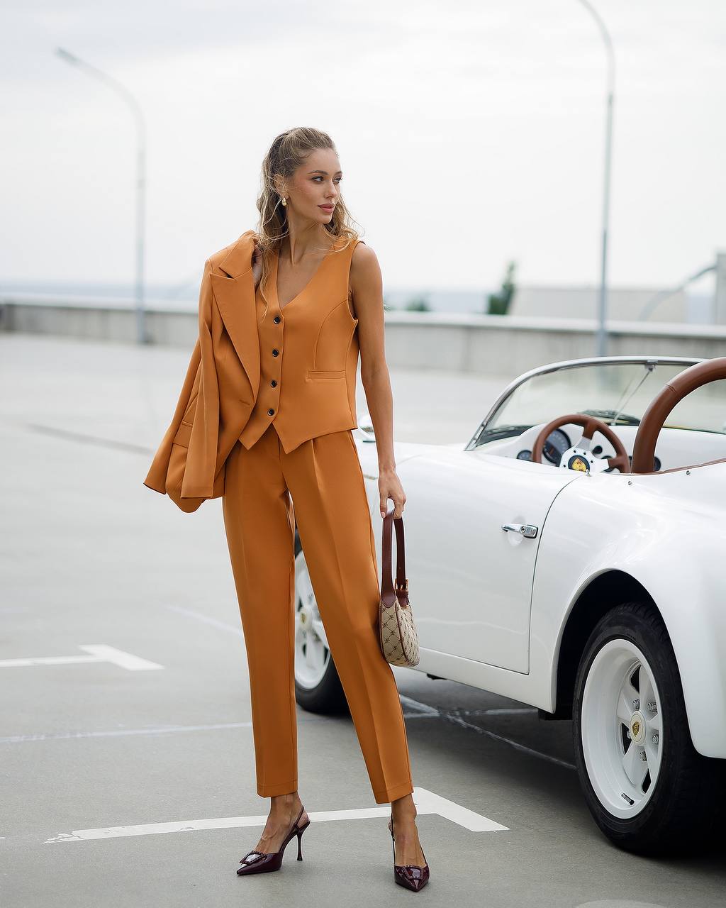 Woman in a mustard yellow suit standing next to a vintage car on a road.