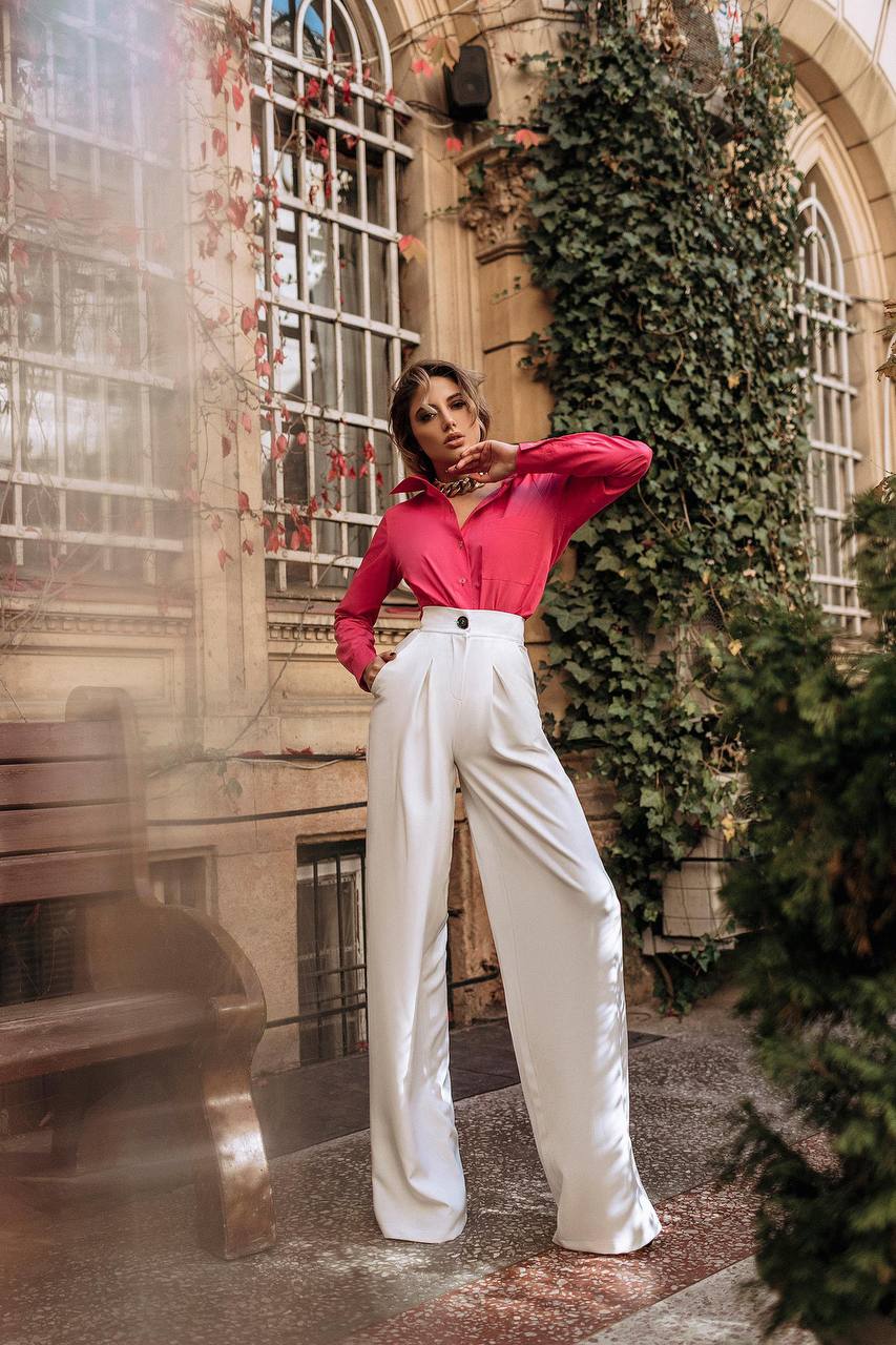 Woman in a red blouse and white pants standing in an outdoor setting with greenery and architectural elements.