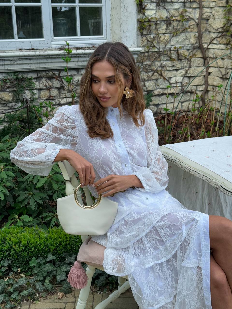 Woman in a white lace dress sitting outdoors holding a cream handbag.