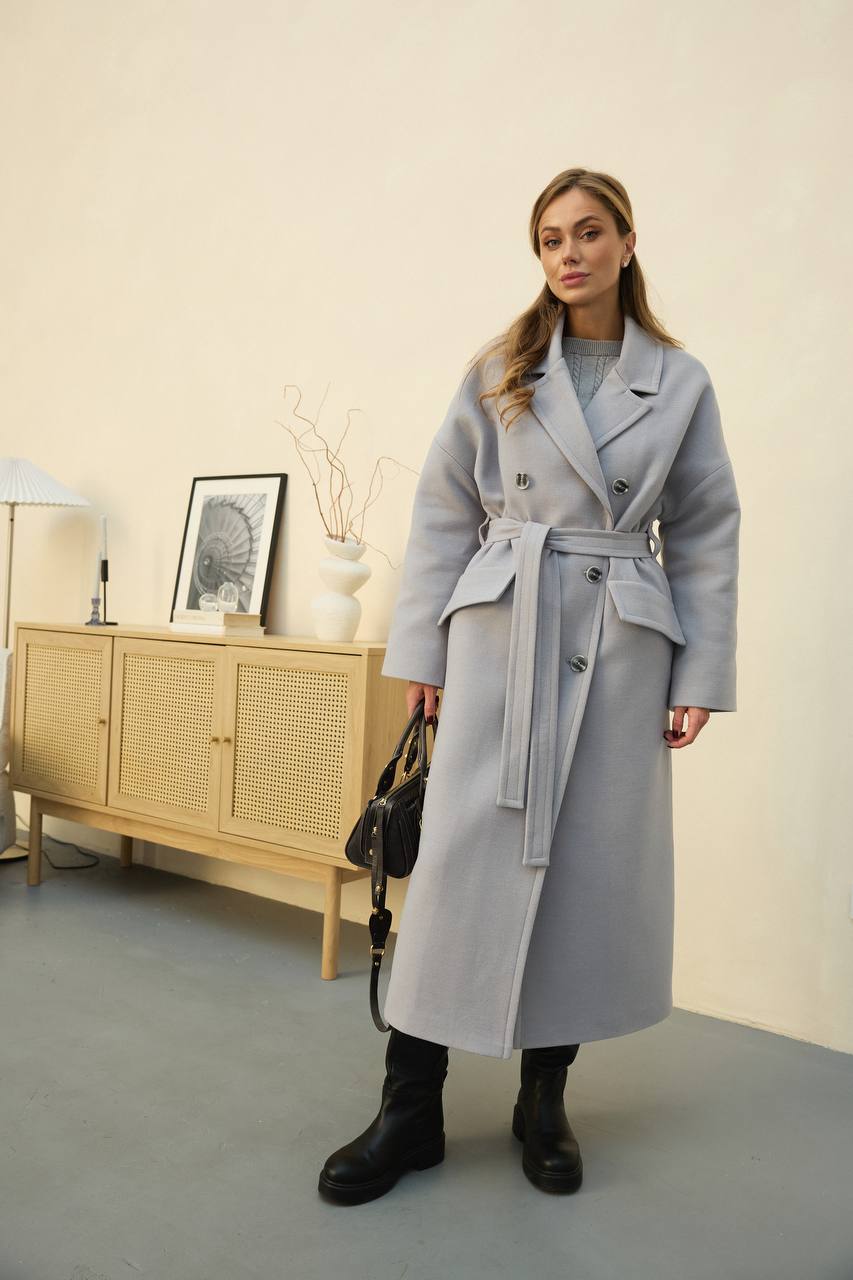Woman wearing a gray coat standing in a room with a light-colored wall and wooden cabinet.
